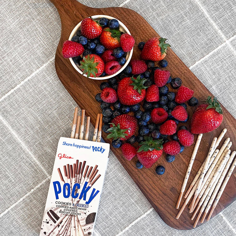 Wooden board with berries, a bowl of berries, and a box of Pocky on a textured surface.